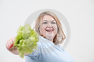 A young lush fat woman in casual blue clothes on a white background holding green lettuce leaves in her hands and mouth.