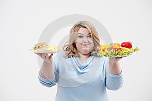 Young lush fat woman in casual blue clothes on a white background holding a vegetable salad and a plate of fast food