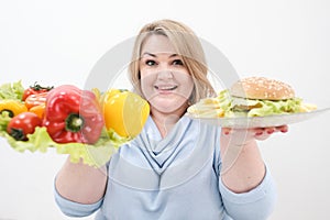 Young lush fat woman in casual blue clothes on a white background holding a vegetable salad and a plate of fast food