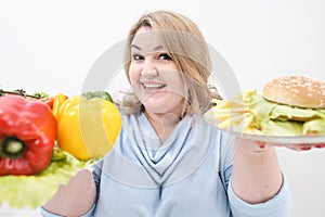 Young lush fat woman in casual blue clothes on a white background holding a vegetable salad and a plate of fast food
