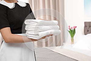 Young maid holding stack of fresh towels in hotel room