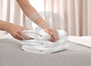 Young maid putting stack of fresh towels on bed in hotel room