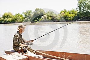 Young male angler holding rod, fishing, while tired dog resting next, breathing hard in boat.