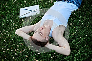 Young male artist  wearing torn jeans and white t-shirt  lying on green grass during sunset  with empty canvas beside  thinking