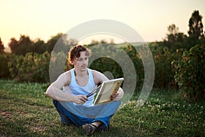 Young male artist, wearing torn jeans and white t-shirt, sitting on ground on green field during sunset, holding empty canvas,