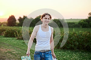 Young male artist  wearing torn jeans and white t-shirt  walking on green field during sunset  holding canvas and palette thinking