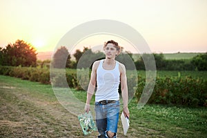 Young male artist  wearing torn jeans and white t-shirt  walking on green field during sunset  holding canvas and palette thinking