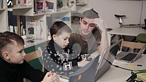 Young male attactive teacher in his 20`s explaining something to two little boys, teaching them how to use laptop