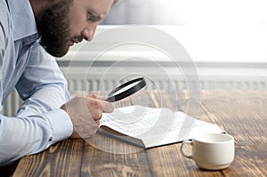 Young male businessman holding a magnifying glass in his hands and reading an open book of the Bible. Sitting at a wooden table