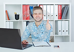 Young male businessman showing OK sign with thumb up sitting at office.