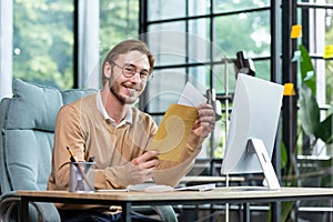 A young male businessman sits in the office, received and holds an envelope with a letter in his hands. Happy smiling at