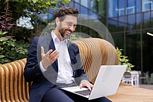 A young male businessman is sitting on a bench near the office wearing headphones, holding a laptop on his lap and