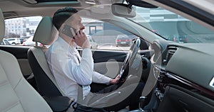 Young male businessman is sitting in the driver's seat in the car and talking on a cell phone. Smiling businessman man