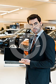 young male car dealer in eyeglasses pointing at laptop with blank screen