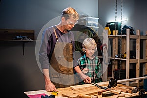 A young male carpenter is teaching woodwork to a young boy in his workshop.