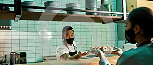 Young male chef of Japanese restaurant wearing protective mask and gloves made sushi rolls, giving them to waitress