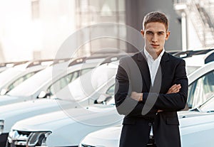 Young male consultant in auto show standing near cars and looking at camera