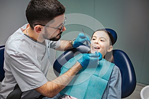 Young male dentist check girl`s teeth. She keep mouth opened. Man use dental tools. Girl sit in chair in room.