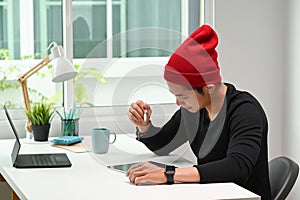 A young male designer in red wool hat working desk with digital graphic tablet and stylus pen.