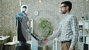 Young male developer and manlike robot cyborg shaking hands standing in office