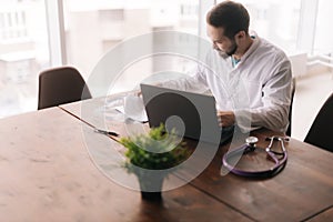 Young male doctor wearing white coat working with patient health data sitting at desk with laptop.