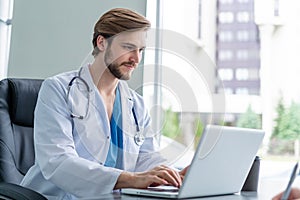 Young male doctor working on laptop computer, sitting in medical office.