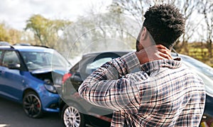 Young Male Driver With Whiplash Injury Standing By Damaged Car After Road Traffic Accident