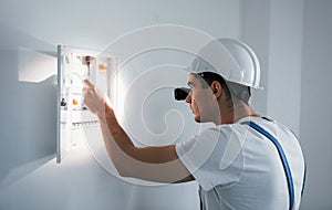 Young male electrician in white hard hat works indoors in the room
