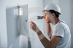 Young male electrician in white hard hat works indoors in the room