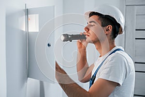 Young male electrician in white hard hat works indoors in the room