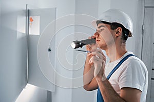 Young male electrician in white hard hat works indoors in the room