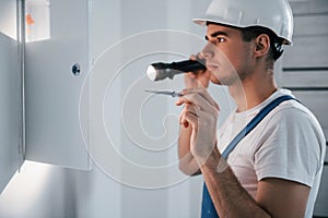 Young male electrician in white hard hat works indoors in the room