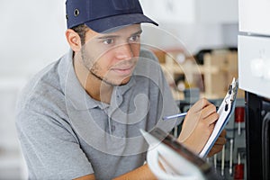 Young male electrician writing on clipboard while looking at oven