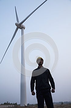 Young male engineer in a hardhat standing in front of a wind turbine at dusk