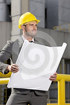 Young male engineer holding blueprint while looking away at construction site