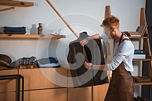 young male fashion designer in apron and eyeglasses looking at suit jacket on mannequin