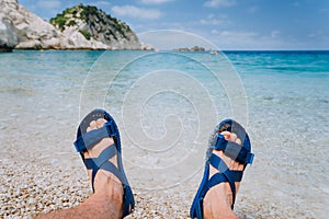 Young male feets wear blue flip-flop sandal sunbathing on pebble beach in front of blue sea water and rocks in