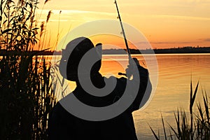 A young male fisherman in a hat throws a fishing rod into the river at sunset. Silhouette of a man in a hat with a fishing rod in