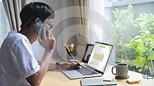 Young male freelancer having cell phone conversation and working with computer laptop a home