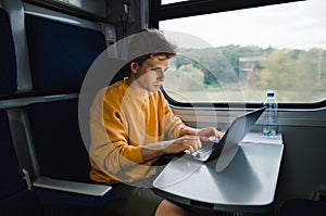 Young male freelancer in orange sweatshirt working on laptop in train with serious face. Work on vacation