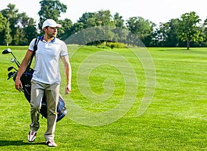 Young Male Golfer carries a Bag of Golf Clubs. Slim Man in White T-shirt , Golf Shoes, White Cap and Beige Pants is