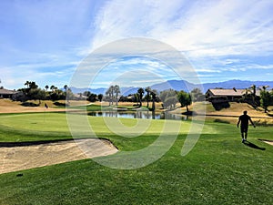 A young male golfer walking towards the green on a par 4 surrounded by water and palm trees in the background on the desert oasis