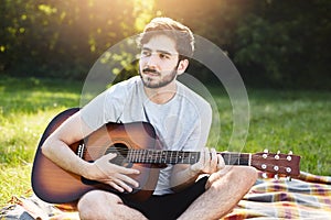 Young male guitarist with trendy hairstyle, dark eyes and thick beard playing guitar at sunset sitting crossed legs at wrap lookin