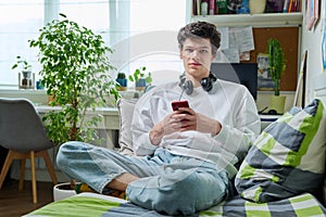 Young male in headphones with phone looking at camera sitting on couch at home