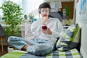 Young male in headphones with phone looking at camera sitting on couch at home