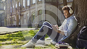 Young male in headphones sitting on grass, leaning against tree, choosing song photo