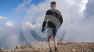 Young male hiker with backpack reaching up top of mountain with cloudy sky at background and raising hands. Boy tourist