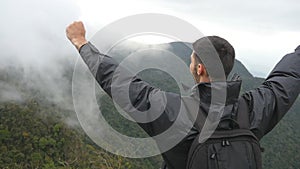 Young male hiker with backpack reaching up top of mountain and raised hands. Man tourist standing on the edge of