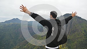 Young male hiker with backpack reaching up top of mountain and raised hands. Man tourist standing on the edge of