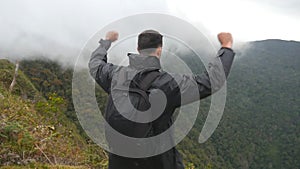 Young male hiker with backpack reaching up top of mountain and raised hands. Man tourist standing on the edge of